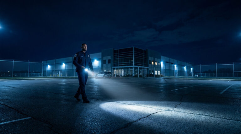 Agent de sécurité en uniforme effectuant une ronde de nuit sur un parking avec une lampe torche.