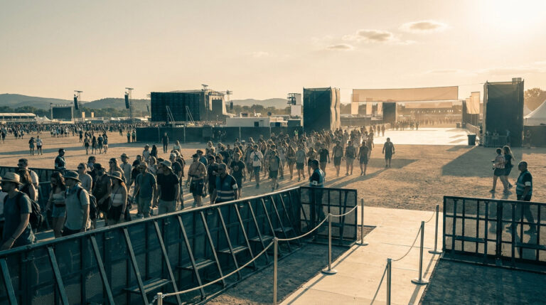 Vue d'un festival en extérieur avec une foule se déplaçant entre des barrières de sécurité sous un soleil couchant.